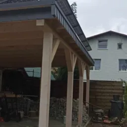 Drewniana konstrukcja z panelami słonecznymi na dachu, widok z boku, w tle dom jednorodzinny i stosy kamieni.