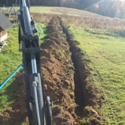 Wykopany wąski rów w ziemi na zielonym terenie, widoczny fragment ramienia koparki z niebieskim przewodem, w tle drzewa i pagórki.