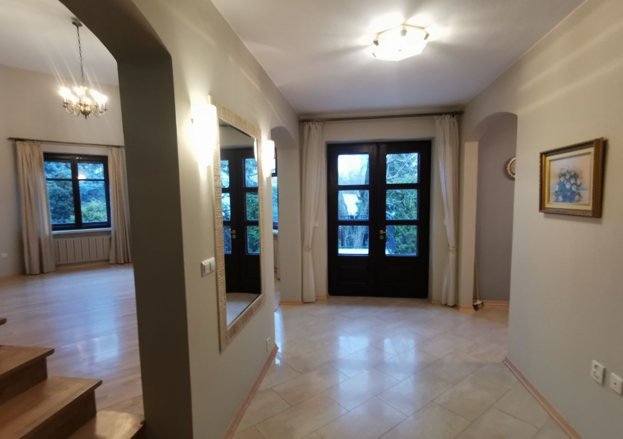 Jasny hol z drewnianymi schodami, marmurową podłogą, lustrem w ozdobnej ramie i oknami z widokiem na zieleń.