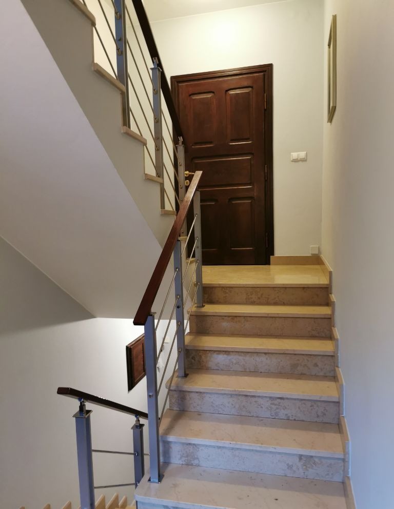 Marmurowe schody z metalową balustradą prowadzące do ciemnych, drewnianych drzwi w jasnym wnętrzu.