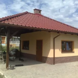 Niewielki budynek z czerwonym dachem i dobudowaną wiatą, wykończony cegłą klinkierową u dołu, z widoczną drabiną opartą o konstrukcję wiaty.