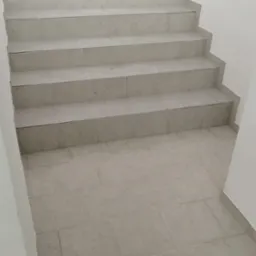 Wykończone schody i podłoga wyłożone jasnymi, kwadratowymi płytkami ceramicznymi, widok z dołu schodów w kierunku góry, białe ściany.