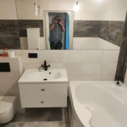 Wykończona łazienka z białą armaturą, czarną baterią umywalkową i wanną, widoczne płytki w dwóch odcieniach oraz odbicie fotografa w lustrze.