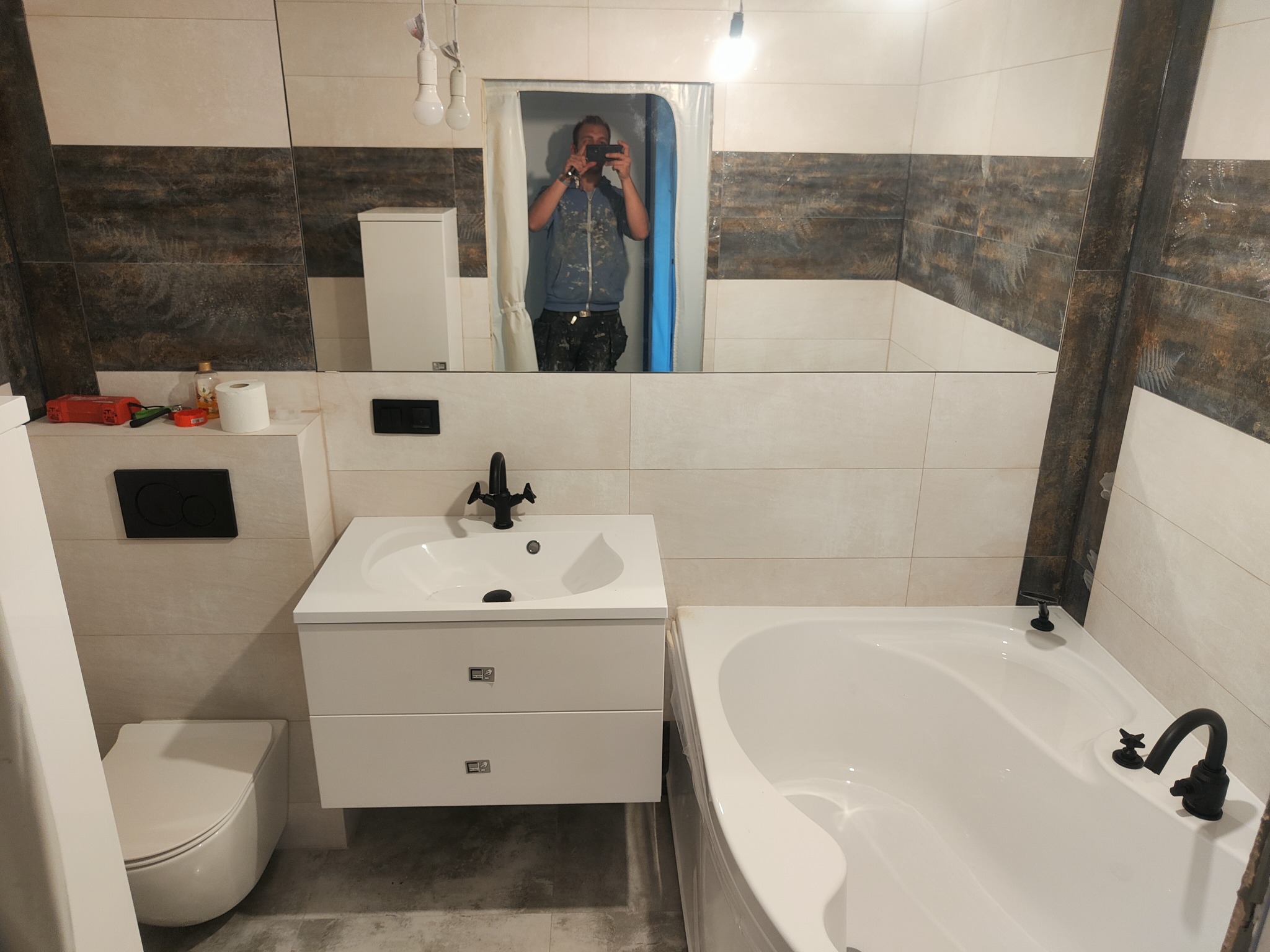 Wykończona łazienka z białą armaturą, czarną baterią umywalkową i wanną, widoczne płytki w dwóch odcieniach oraz odbicie fotografa w lustrze.