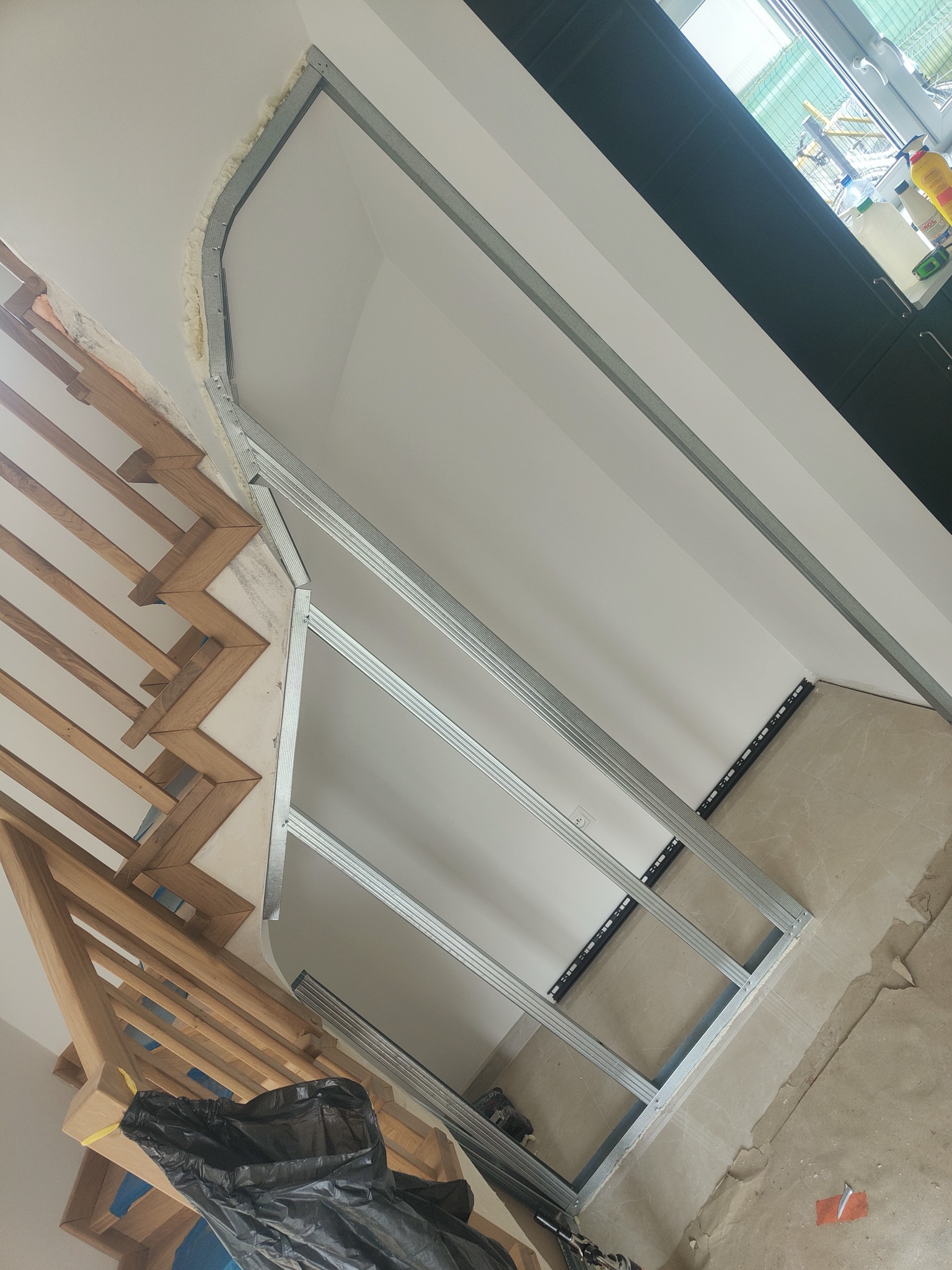 Metalowy stelaż pod zabudowę wnęki schodowej, wypełniony pianką montażową, widoczne drewniane schody i fragment podłogi z płytek.