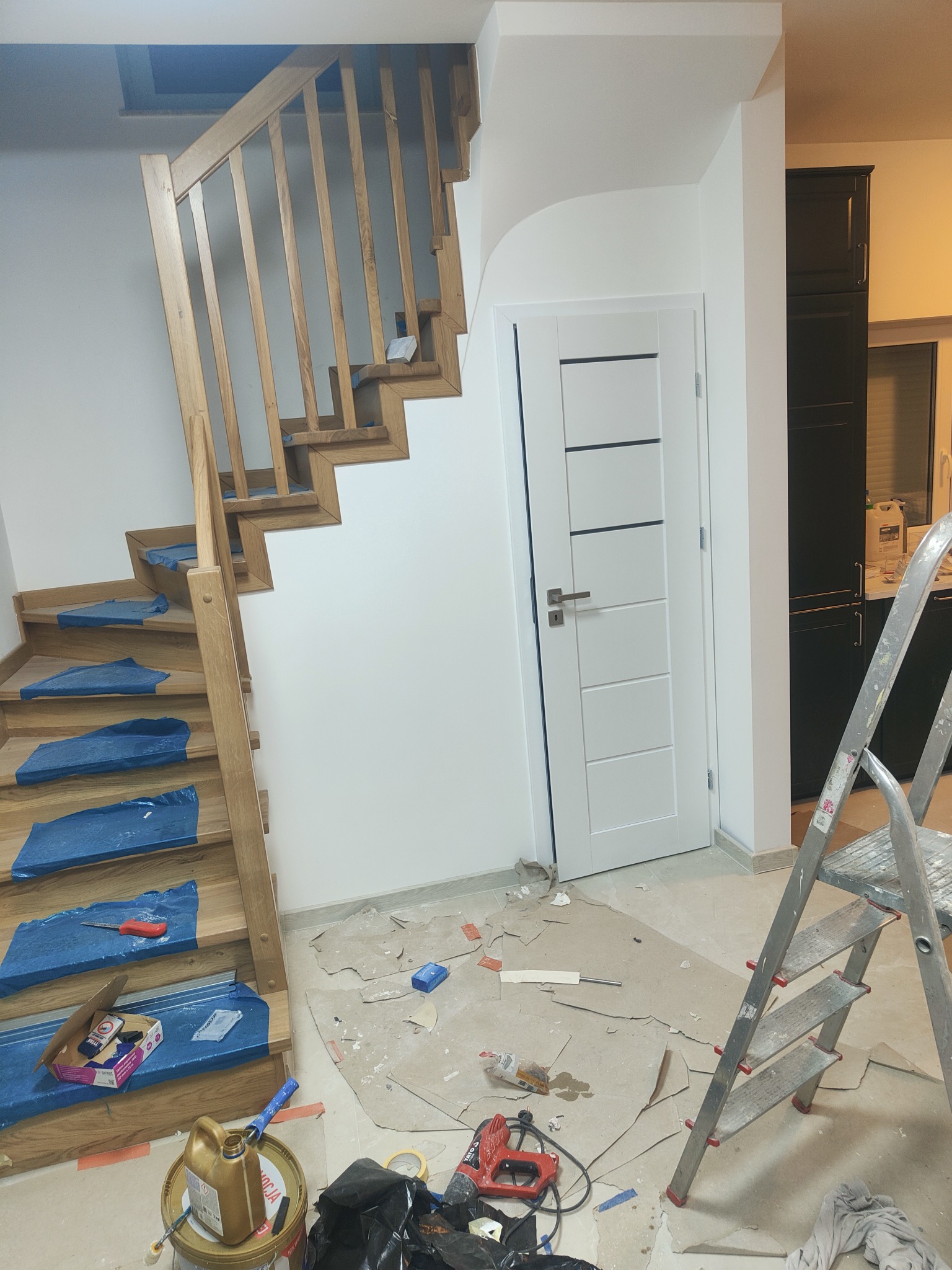 Klatka schodowa z drewnianymi schodami zabezpieczonymi folią, białe ściany po malowaniu, widoczne drobne zabrudzenia i kartony ochronne na podłodze oraz drabina aluminiowa. W tle białe drzwi...
