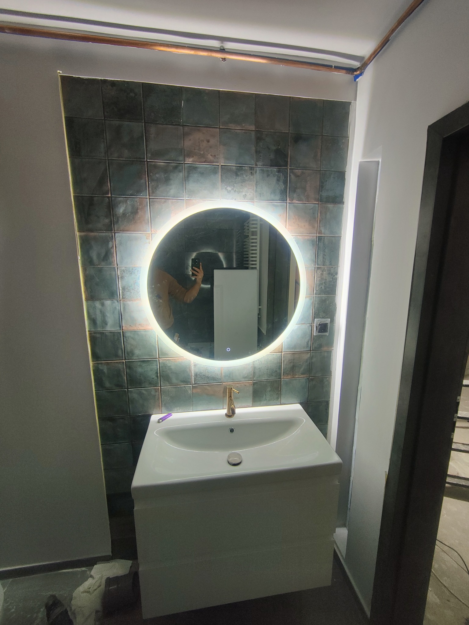 Łazienka z umywalką nablatową, baterią w kolorze złotym i okrągłym lustrem z podświetleniem LED na tle ściany wyłożonej ciemnymi, kwadratowymi płytkami z efektem postarzenia.