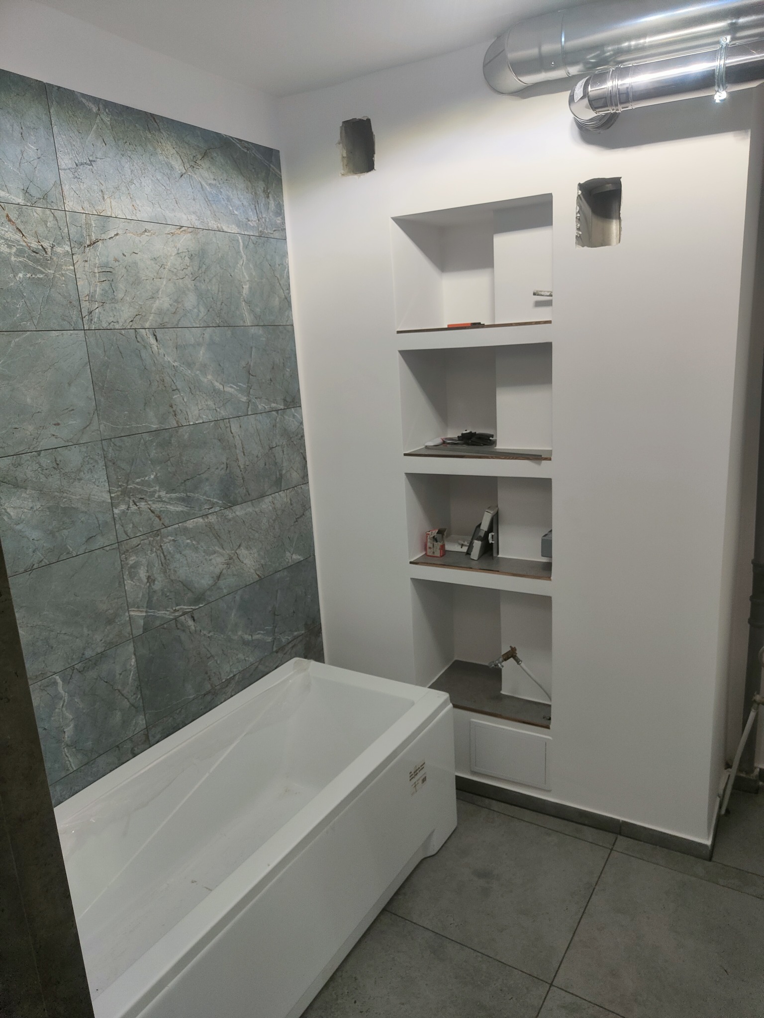 Wnętrze łazienki w trakcie remontu: biała wanna z folią ochronną, ściana wyłożona szarymi płytkami imitującymi marmur, wnęka w ścianie z półkami, widoczne instalacje i otwory w ścianie.