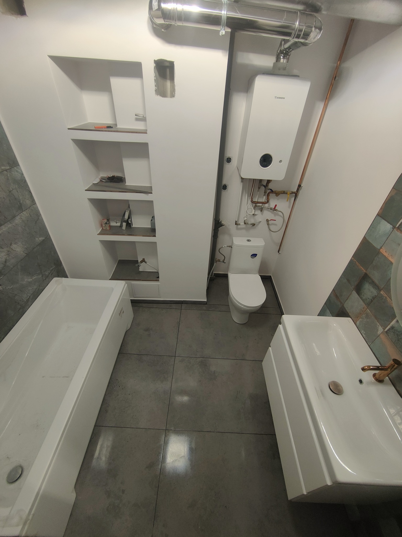 Wykończona łazienka z białą wanną, umywalką na postumencie z mosiężną armaturą, toaletą, piecem gazowym Junkers i wnękami w ścianie.