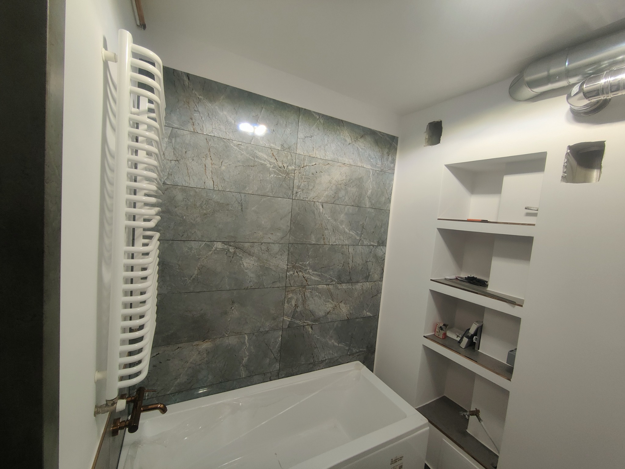 Wykończona łazienka z wanną zabezpieczoną folią, grzejnikiem drabinkowym z miedzianą armaturą, szarą ścianą z płytek imitujących marmur oraz wnęką z półkami.