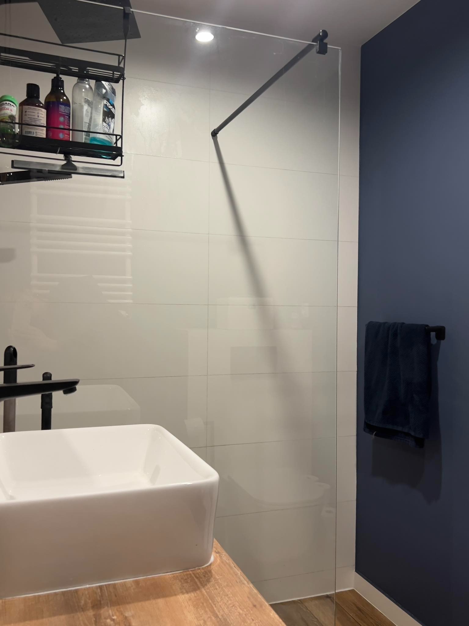 Nowoczesna łazienka z białą umywalką nablatową, szklaną kabiną prysznicową z czarnymi elementami i granatową ścianą z ręcznikiem.