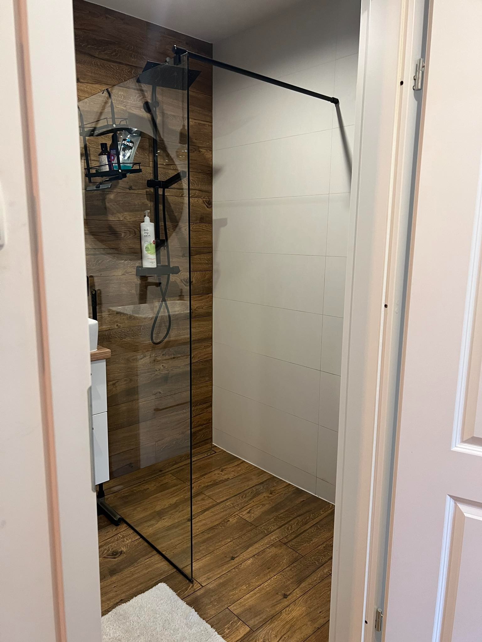 Nowoczesna kabina prysznicowa z czarną armaturą, drewnopodobnymi płytkami na ścianie i podłodze, widok zza uchylonych drzwi.