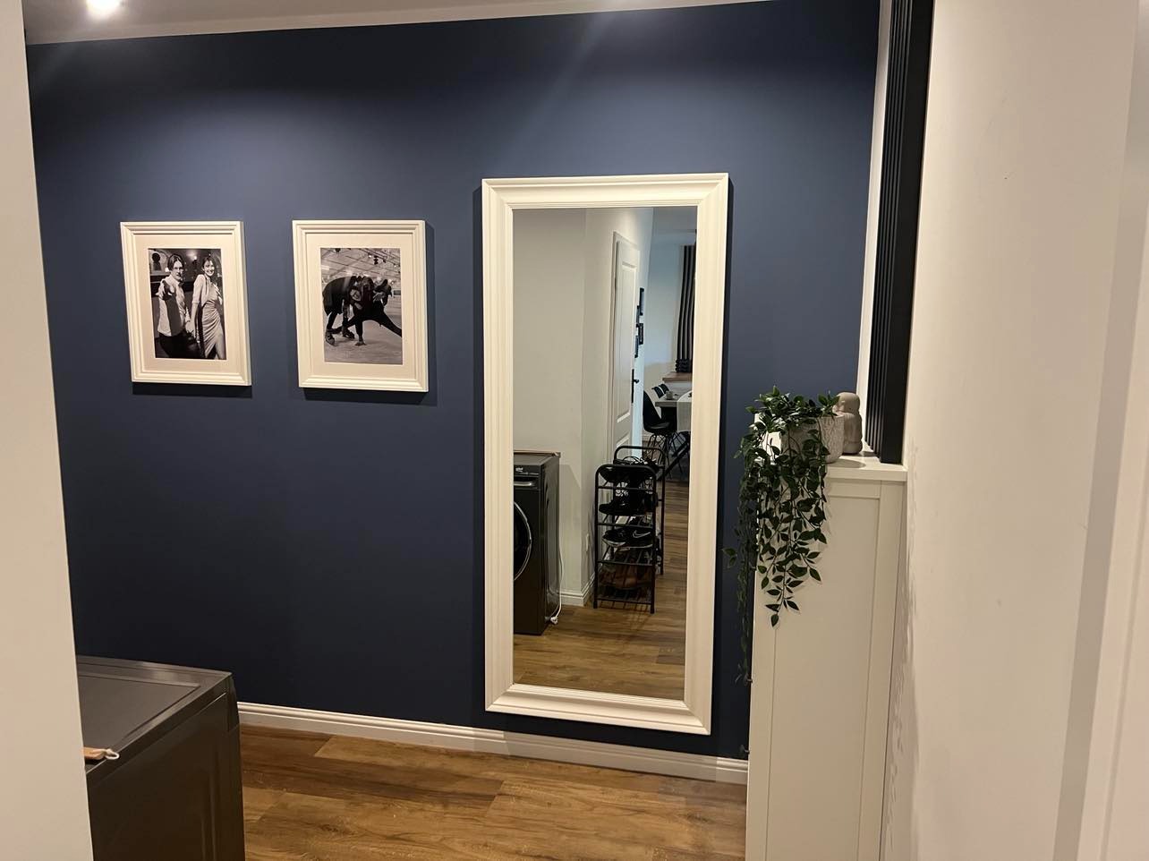 Granatowa ściana z dwoma czarno-białymi zdjęciami w białych ramkach, duże lustro w białej ramie odbijające widok na wnętrze mieszkania, drewniana podłoga i biała szafka z rośliną.
