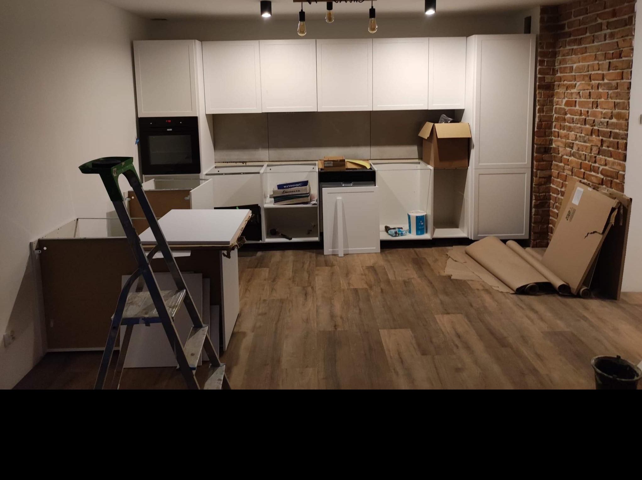 Montaż białych szafek kuchennych w trakcie realizacji, widoczna drabina, kartony i zabezpieczenia podłogi, ściana z cegły w tle.