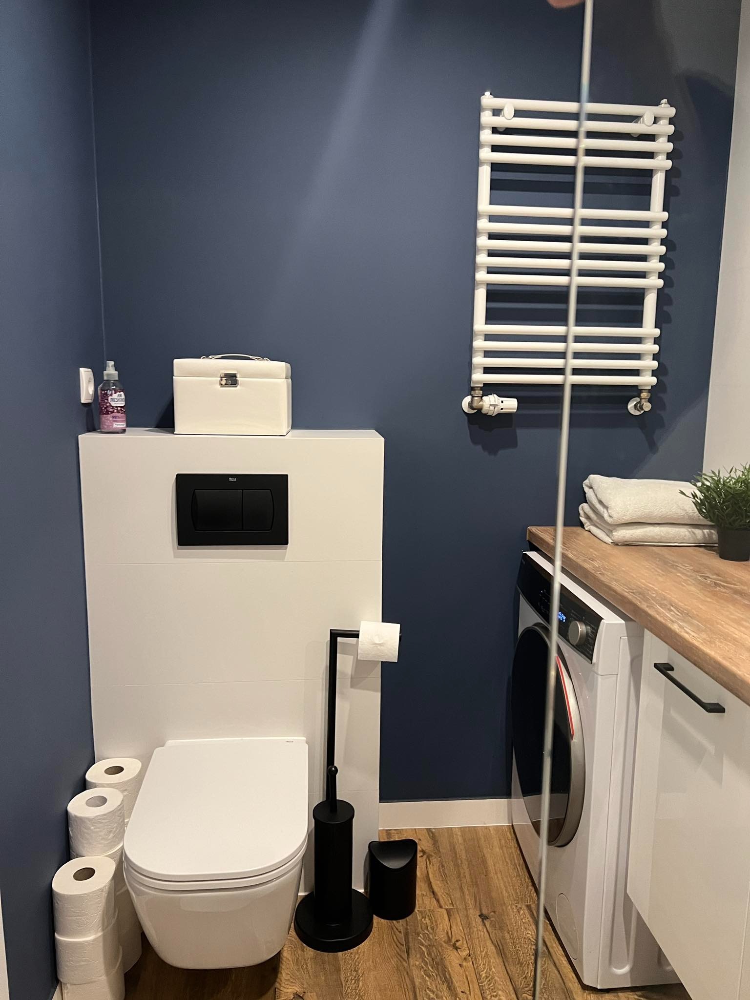 Nowoczesna łazienka w odcieniach bieli i granatu z podwieszaną toaletą, grzejnikiem drabinkowym, pralką pod blatem z drewnianym wykończeniem i dekoracyjnymi akcentami.