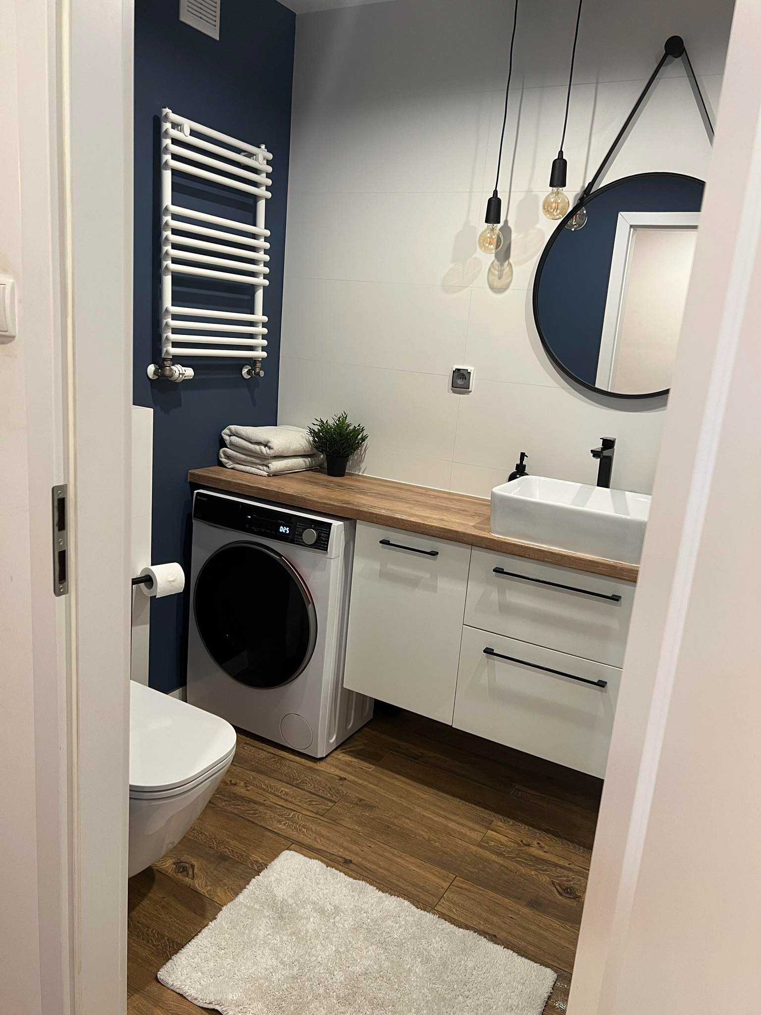 Nowoczesna łazienka z pralką pod blatem, okrągłym lustrem i drewnianą podłogą, widok zza drzwi.
