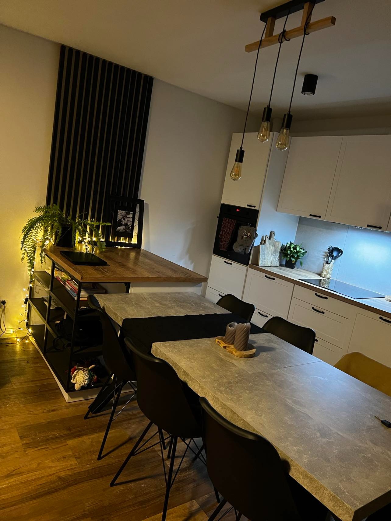 Nowoczesna kuchnia z białymi szafkami, drewnianym blatem roboczym, stołem z blatem imitującym beton i czarnymi krzesłami, oświetlona trzema wiszącymi lampami z żarówkami Edisona i dodatkowym...