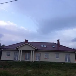 Nowy dom jednorodzinny z czerwoną dachówką, widoczne okna dachowe i komin, elewacja w trakcie wykończenia, okna zabezpieczone folią, ujęcie z poziomu gruntu.