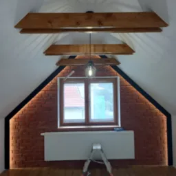 Poddasze po remoncie: ściana z cegły klinkierowej z oświetleniem LED, drewniane belki stropowe, lampa wisząca, grzejnik, drabina i wentylator pod oknem.