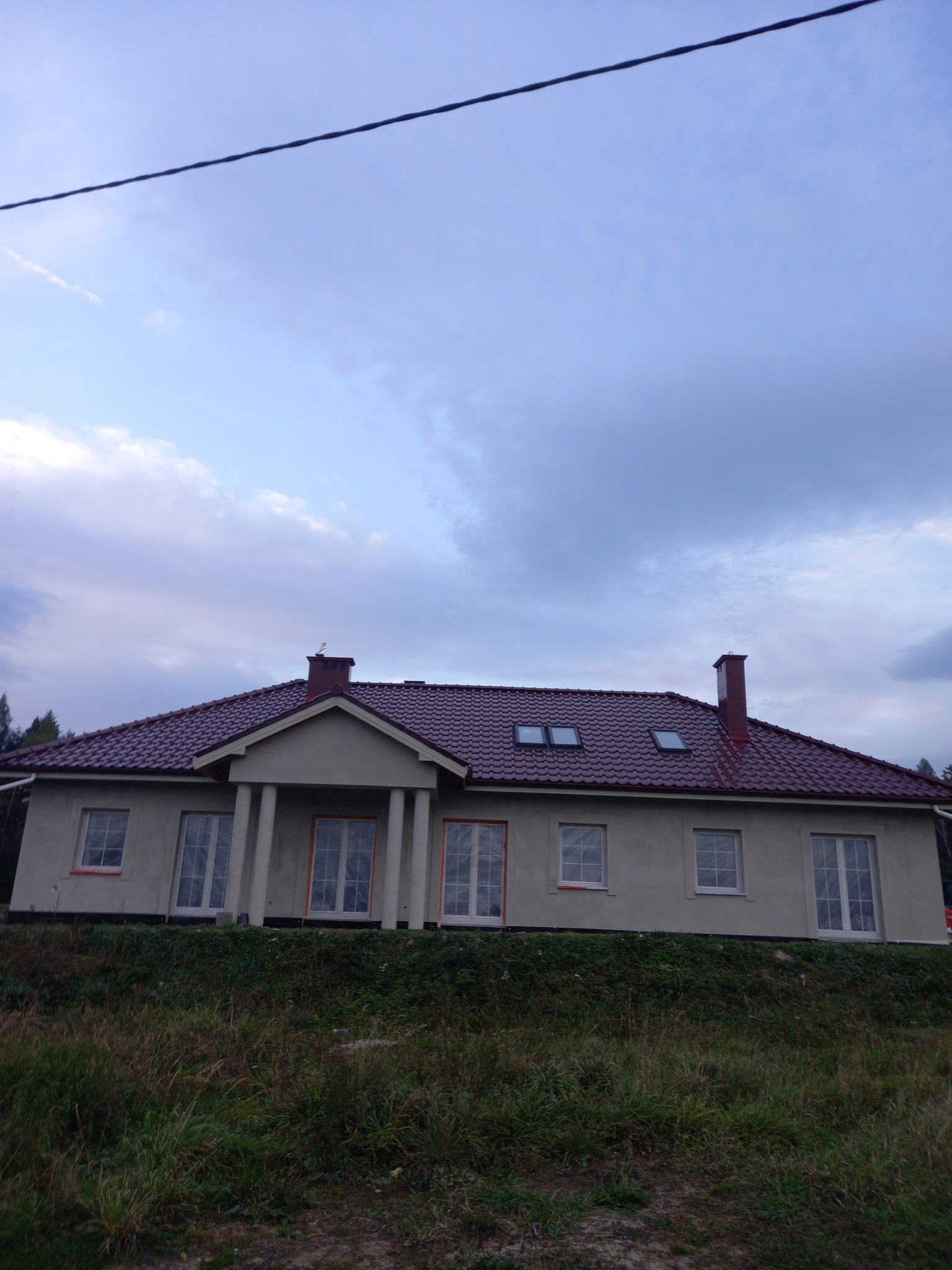 Nowy dom jednorodzinny z czerwoną dachówką, widoczne okna dachowe i komin, elewacja w trakcie wykończenia, okna zabezpieczone folią, ujęcie z poziomu gruntu.