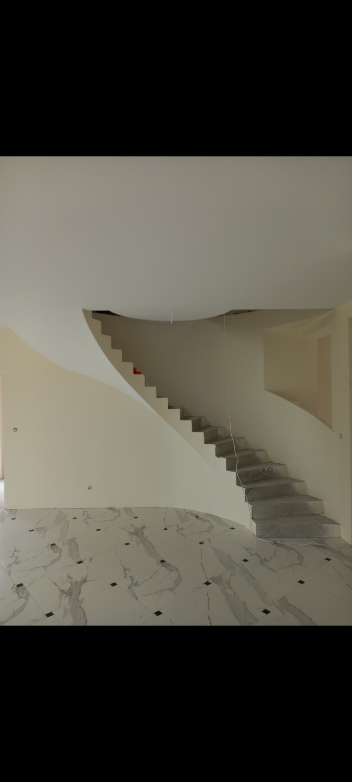 Wnętrze w trakcie wykańczania: widoczne surowe schody betonowe, świeżo wyszpachlowane ściany w odcieniach beżu oraz marmurowe płytki podłogowe z czarnymi akcentami.