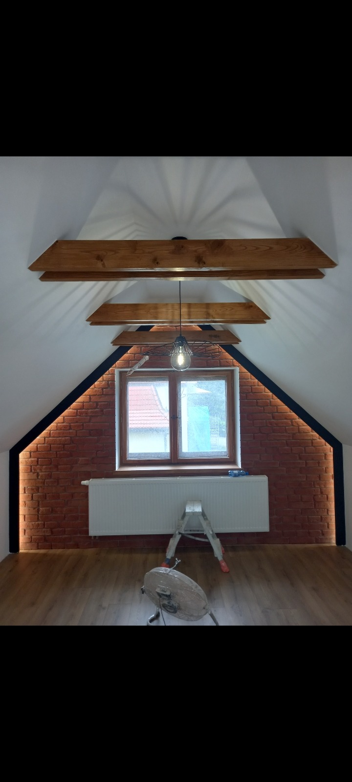 Poddasze po remoncie: ściana z cegły klinkierowej z oświetleniem LED, drewniane belki stropowe, lampa wisząca, grzejnik, drabina i wentylator pod oknem.