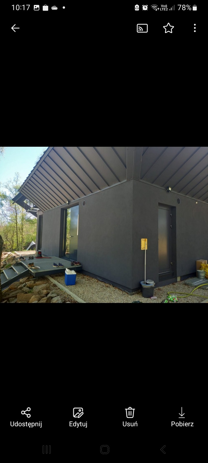 Nowoczesny budynek z szarą elewacją i metalowym dachem, widoczne drzwi wejściowe, okno, teren wokół budynku z kamieniami i narzędziami budowlanymi.