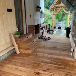 Zadaszony taras z drewnianą podłogą, schodami i obudową z kamienia, na którym leży pies, w tle murowany grill i altana z bali