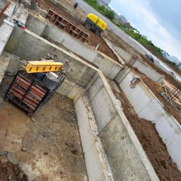 Scand Pref Hause - Budowa fundamentów w Iwkowej: Wykopane i zalane betonem rowy fundamentowe, widoczne zbrojenia i maszyna budowlana w tle, ziemia częściowo zasypana.