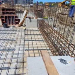 Zbrojenie fundamentu z widocznymi prętami stalowymi, betonowymi elementami i szalunkami, na tle placu budowy z pracownikami i maszynami.