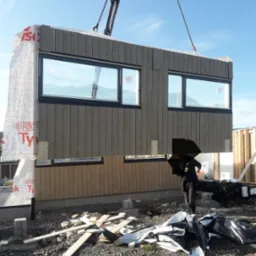 Montaż segmentu domu z drewnianą elewacją i oknami, podnoszonego przez dźwig na placu budowy w słoneczny dzień.