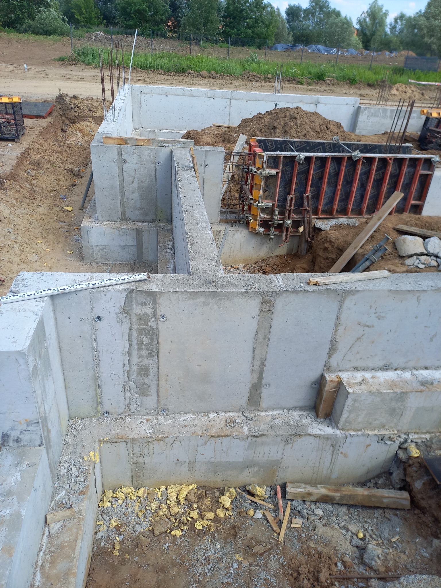 Wylane betonowe fundamenty domu jednorodzinnego, widoczne szalunki, zbrojenia i prace przygotowawcze do dalszych etapów budowy, w tle teren budowy z zielenią.