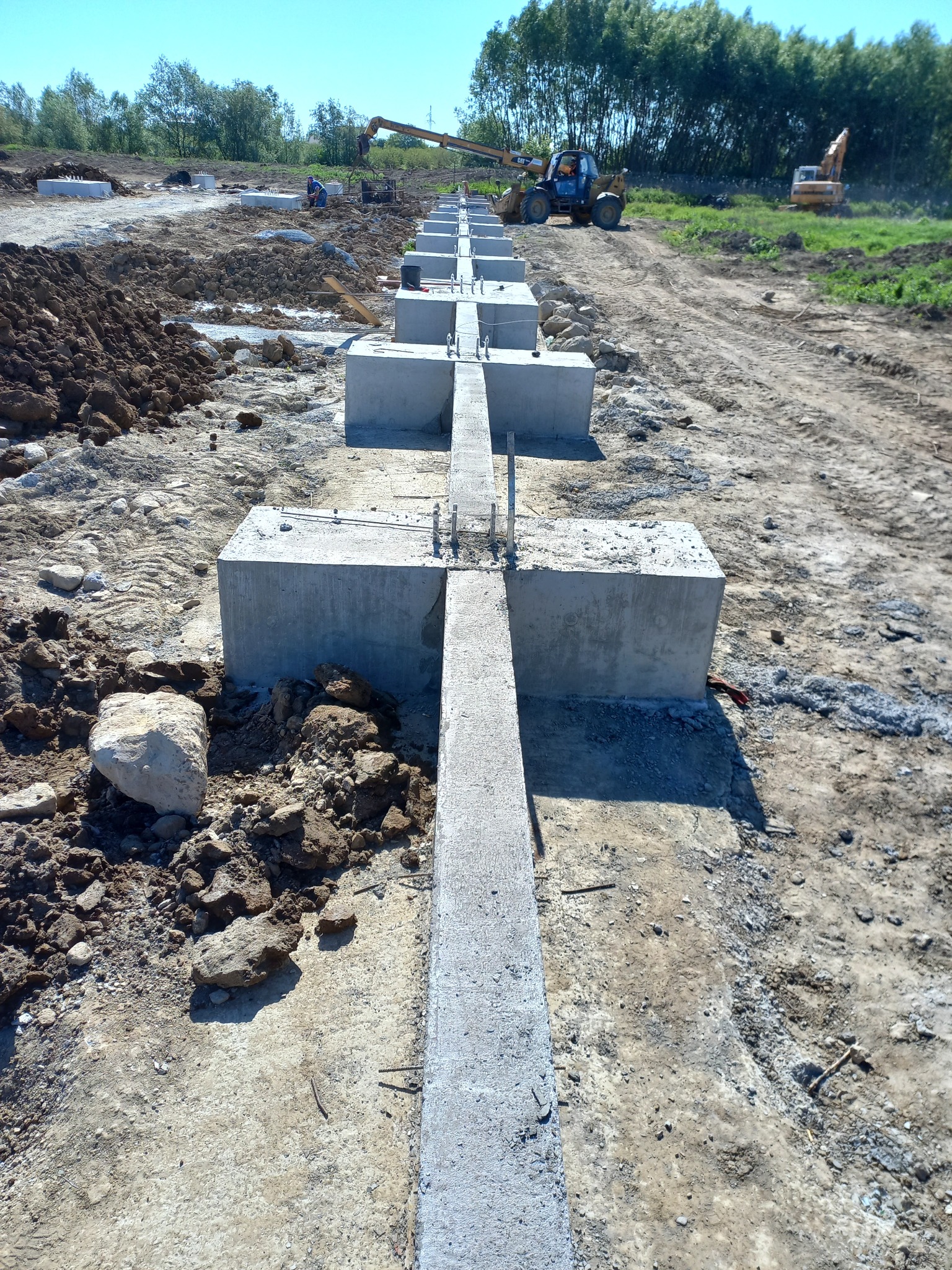Wykop pod budowę z ułożonymi betonowymi fundamentami w kształcie krzyża, widoczny ciężki sprzęt budowlany w tle, ziemia i gruz wokół.