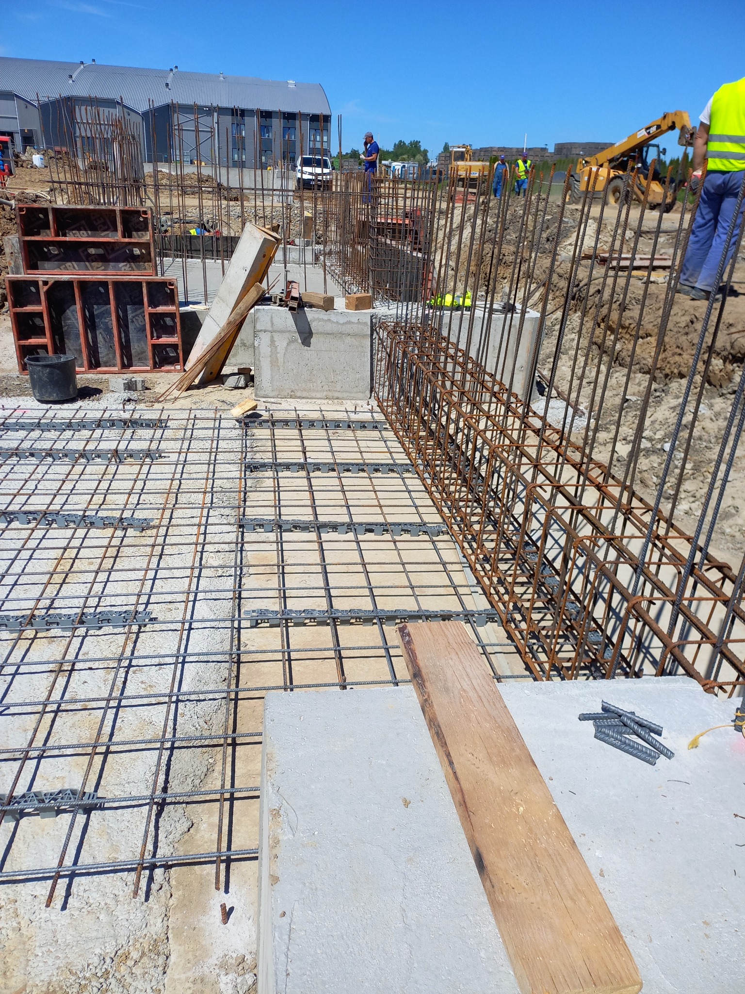 Zbrojenie fundamentu z widocznymi prętami stalowymi, betonowymi elementami i szalunkami, na tle placu budowy z pracownikami i maszynami.