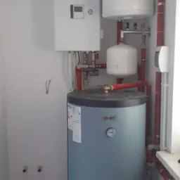 Zamknięte pomieszczenie z zainstalowanym bojlerem Vosti, zbiornikiem wyrównawczym, oraz piecem grzewczym Toshiba, połączonymi czerwoną instalacją hydrauliczną.
