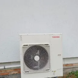 Zewnętrzna jednostka klimatyzacji Toshiba na białym stelażu, ustawiona przy ścianie budynku z widoczną ceglaną podstawą i zielenią.