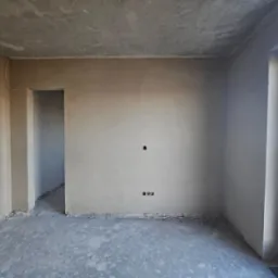 Wnętrze pokoju w stanie deweloperskim po nałożeniu tynków, widoczne gładkie ściany, surowy beton na podłodze oraz otwory na gniazdka elektryczne.