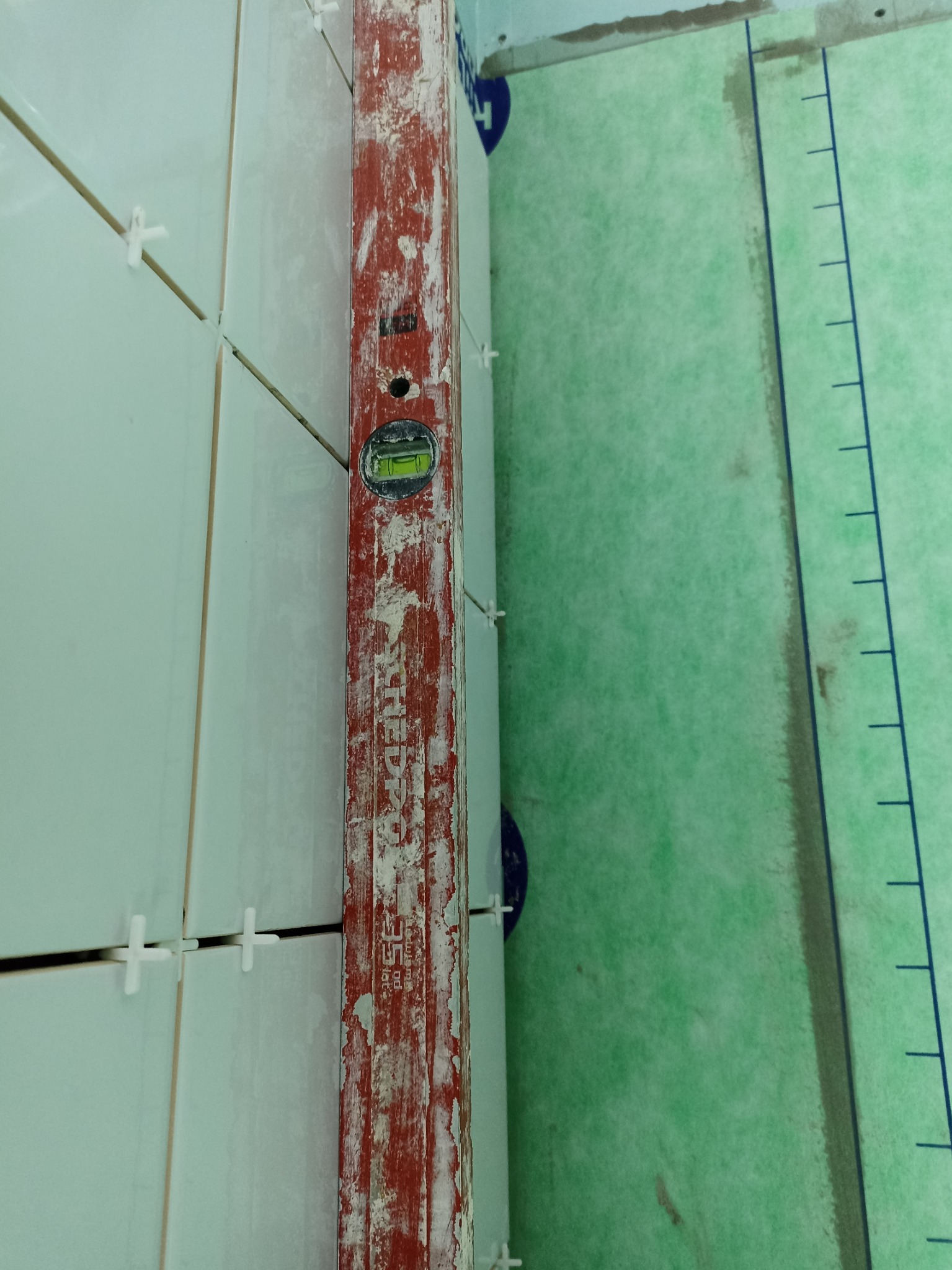 Świeżo ułożone, białe płytki ścienne z plastikowymi krzyżykami dystansowymi, sprawdzane poziomnicą w kolorze czerwonym z widocznymi śladami zaprawy, na ścianie pokrytej zieloną folią z nadrukowaną...