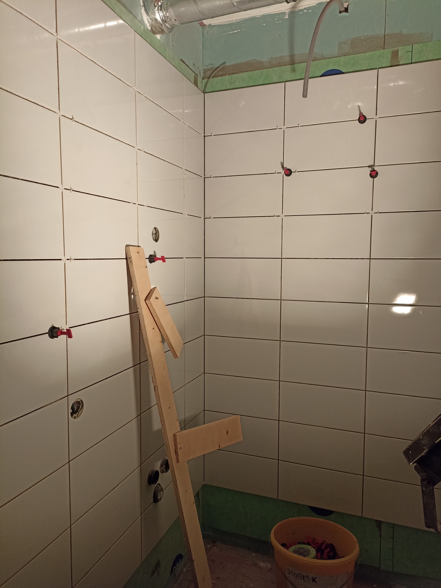 Świeżo ułożone, białe płytki ceramiczne na ścianach w łazience, z widocznymi krzyżykami dystansowymi i czerwonymi elementami poziomującymi, drewniana konstrukcja pomocnicza oparta o ścianę...