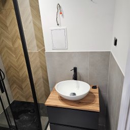 EUROBUD REMONTY WYKOŃCZENIOWE MYKOLA MUKIIENKO - Nowoczesna łazienka z kabiną prysznicową typu walk-in z czarnymi profilami, umywalką nablatową na szafce z drewnianym blatem, szare płytki na ścianach i podłodze, widoczne niedokończone podłączenia...