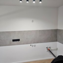 EUROBUD REMONTY WYKOŃCZENIOWE MYKOLA MUKIIENKO - Ściana w trakcie wykańczania z szarymi płytkami, widoczne gniazdka elektryczne i instalacja wodno-kanalizacyjna, oświetlenie sufitowe punktowe.