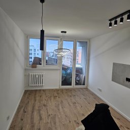 EUROBUD REMONTY WYKOŃCZENIOWE MYKOLA MUKIIENKO - Pusty pokój z drewnianą podłogą, białymi ścianami, grzejnikiem pod oknem balkonowym, nowoczesnym oświetleniem sufitowym i czarną kurtką na krześle.