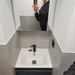 EUROBUD REMONTY WYKOŃCZENIOWE MYKOLA MUKIIENKO - Nowoczesna łazienka po remoncie: biała umywalka z czarną armaturą i szafką w kolorze antracytu, szare płytki na ścianach, odbicie fotografa w lustrze.