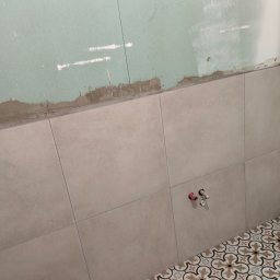 EUROBUD REMONTY WYKOŃCZENIOWE MYKOLA MUKIIENKO - Fragment ściany łazienki z częściowo położonymi beżowymi płytkami na ścianie i dekoracyjną mozaiką na podłodze; widoczne wystające rury instalacji wodnej i surowa ściana nad płytkami, przygotowana...