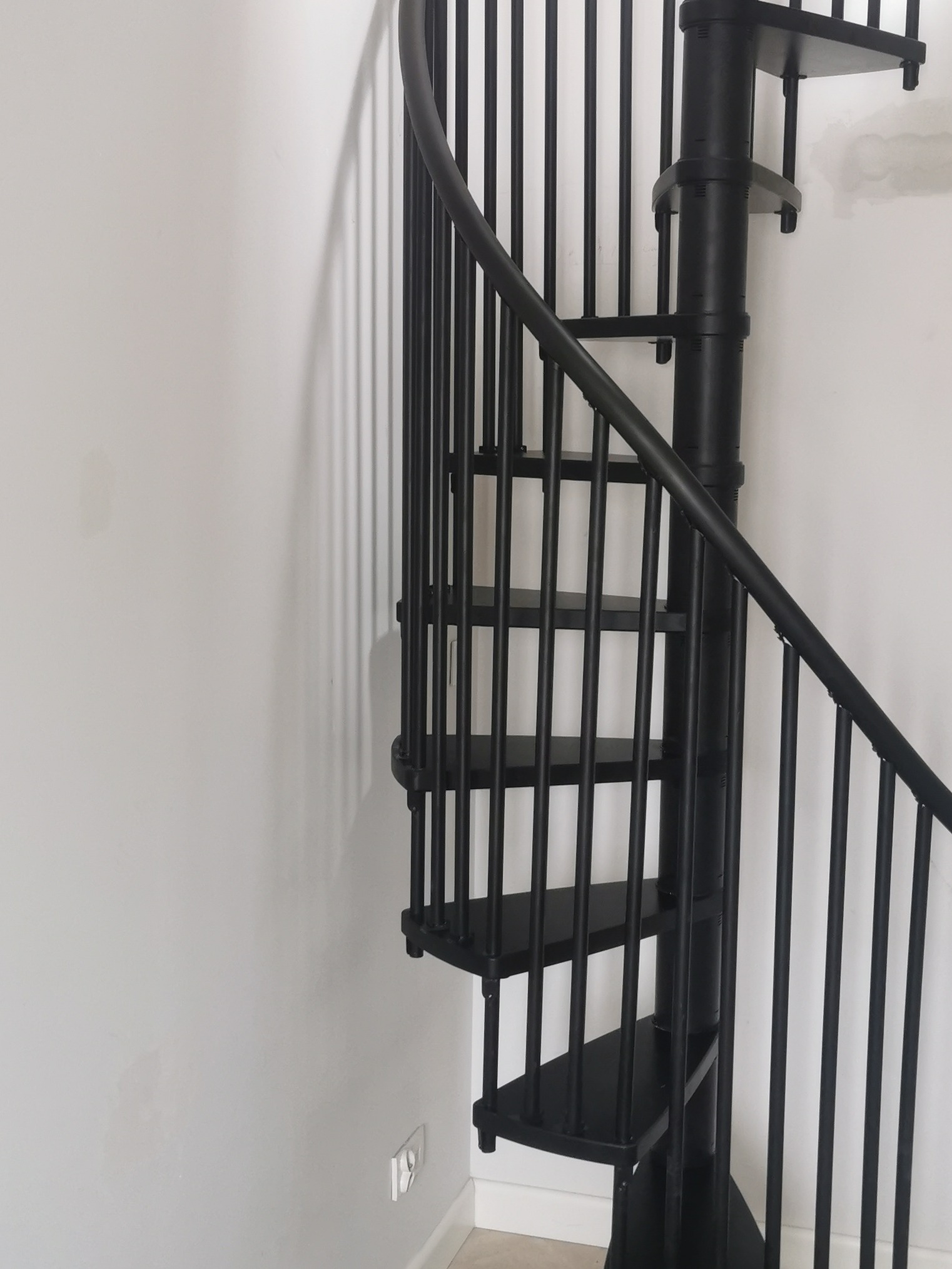 Czarne, metalowe schody spiralne z poręczą, umieszczone przy białej ścianie z widocznym gniazdkiem elektrycznym.