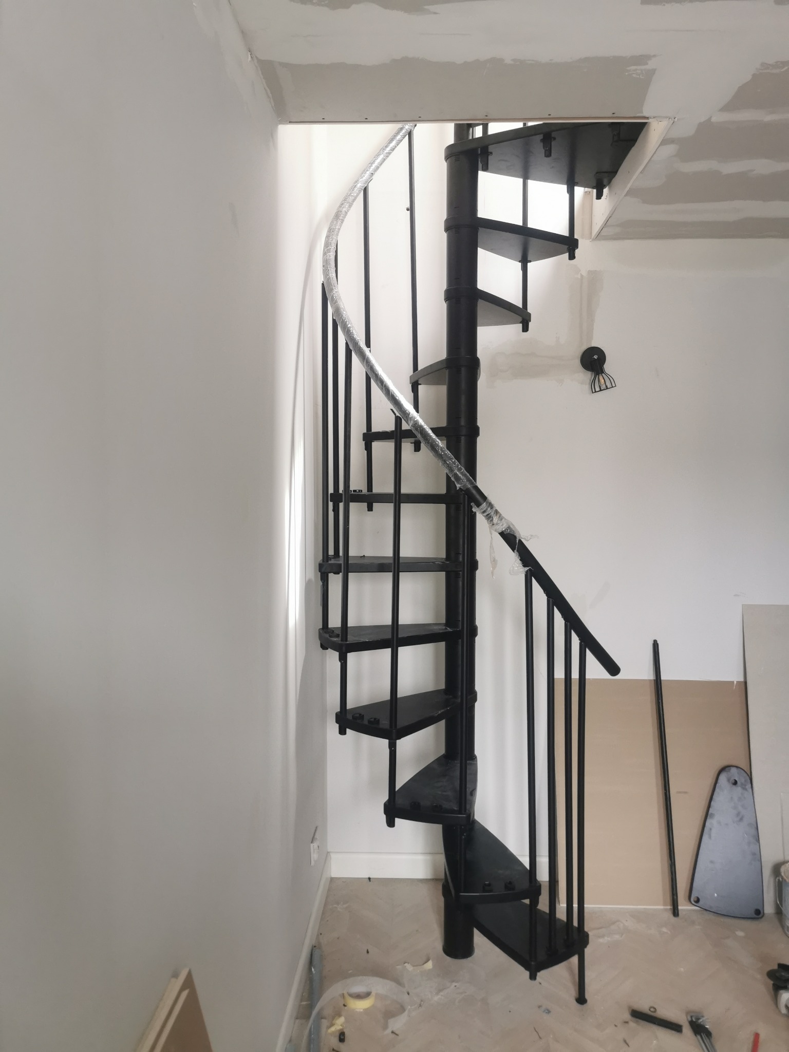 Czarna, metalowa klatka schodowa spiralna w trakcie montażu, zabezpieczona folią, na tle białych ścian i surowego sufitu z płyt kartonowo-gipsowych.
