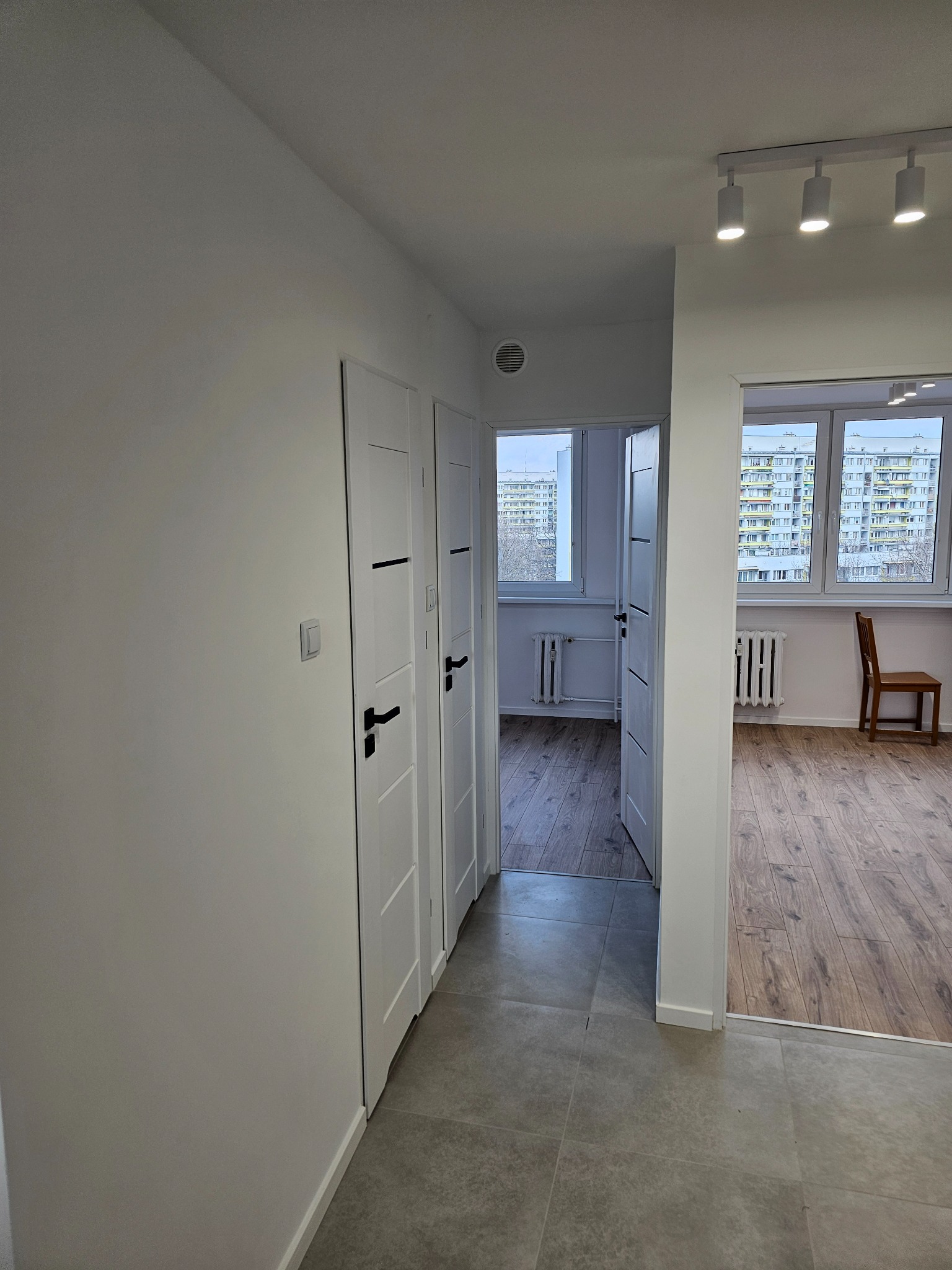 Korytarz w mieszkaniu po odświeżeniu ścian, z trzema białymi drzwiami z czarnymi klamkami, szarą podłogą z płytek i widokiem na pokoje z panelami podłogowymi oraz grzejnikami pod oknami...