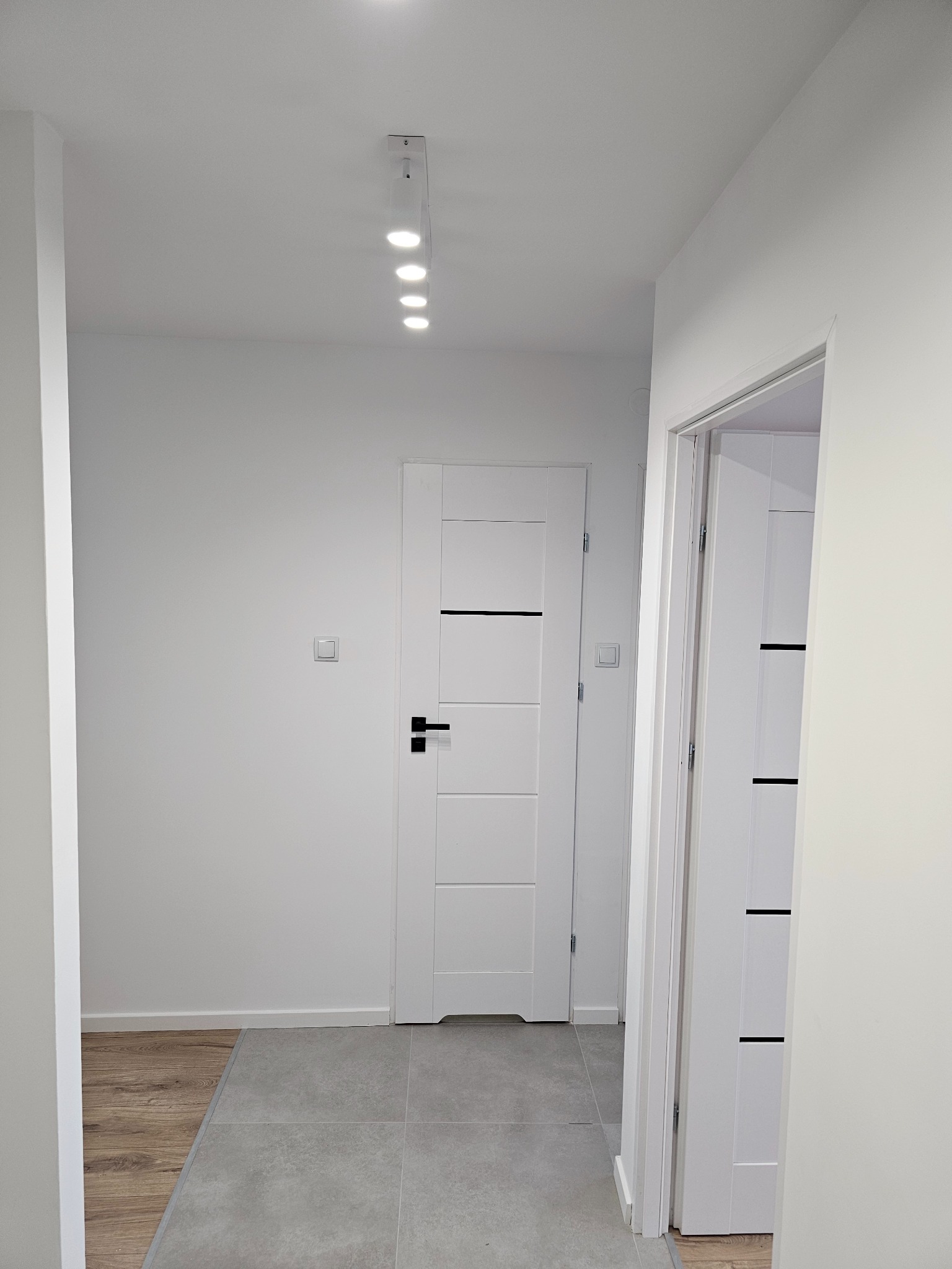 Nowoczesny korytarz z białymi ścianami, szarą podłogą z płytek i panelami, białymi drzwiami z czarnymi akcentami oraz listwą sufitową z oświetleniem punktowym.