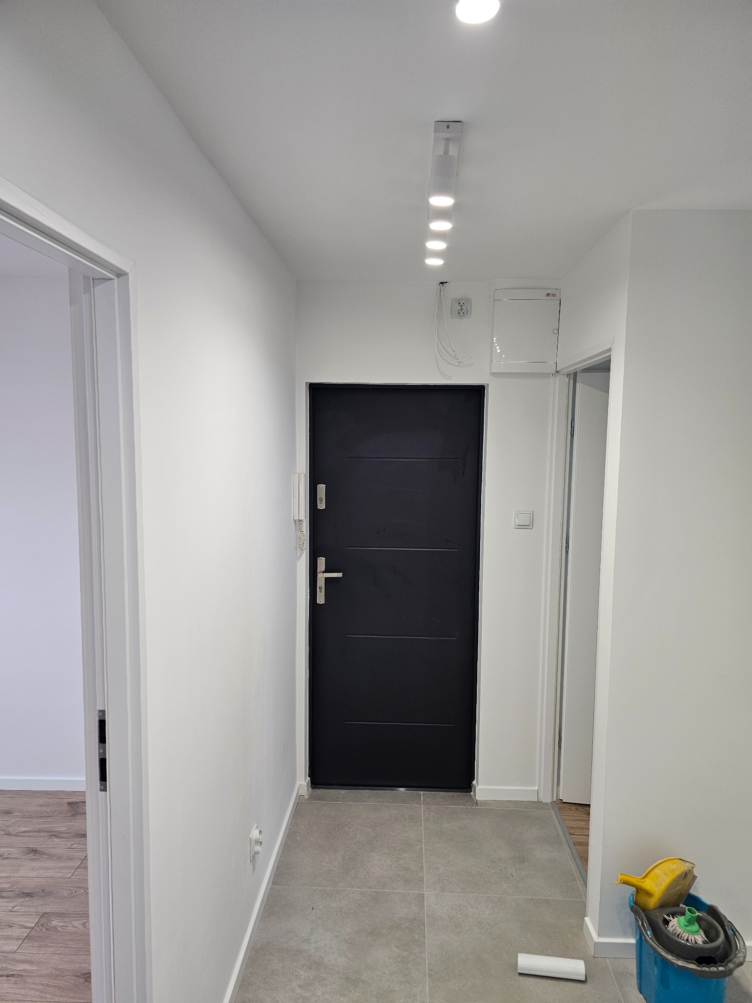 Wnętrze korytarza po odświeżeniu: białe ściany, szara podłoga z płytek, czarne drzwi wejściowe, oświetlenie sufitowe, widoczne ślady prac wykończeniowych - wiadro i mop w rogu.