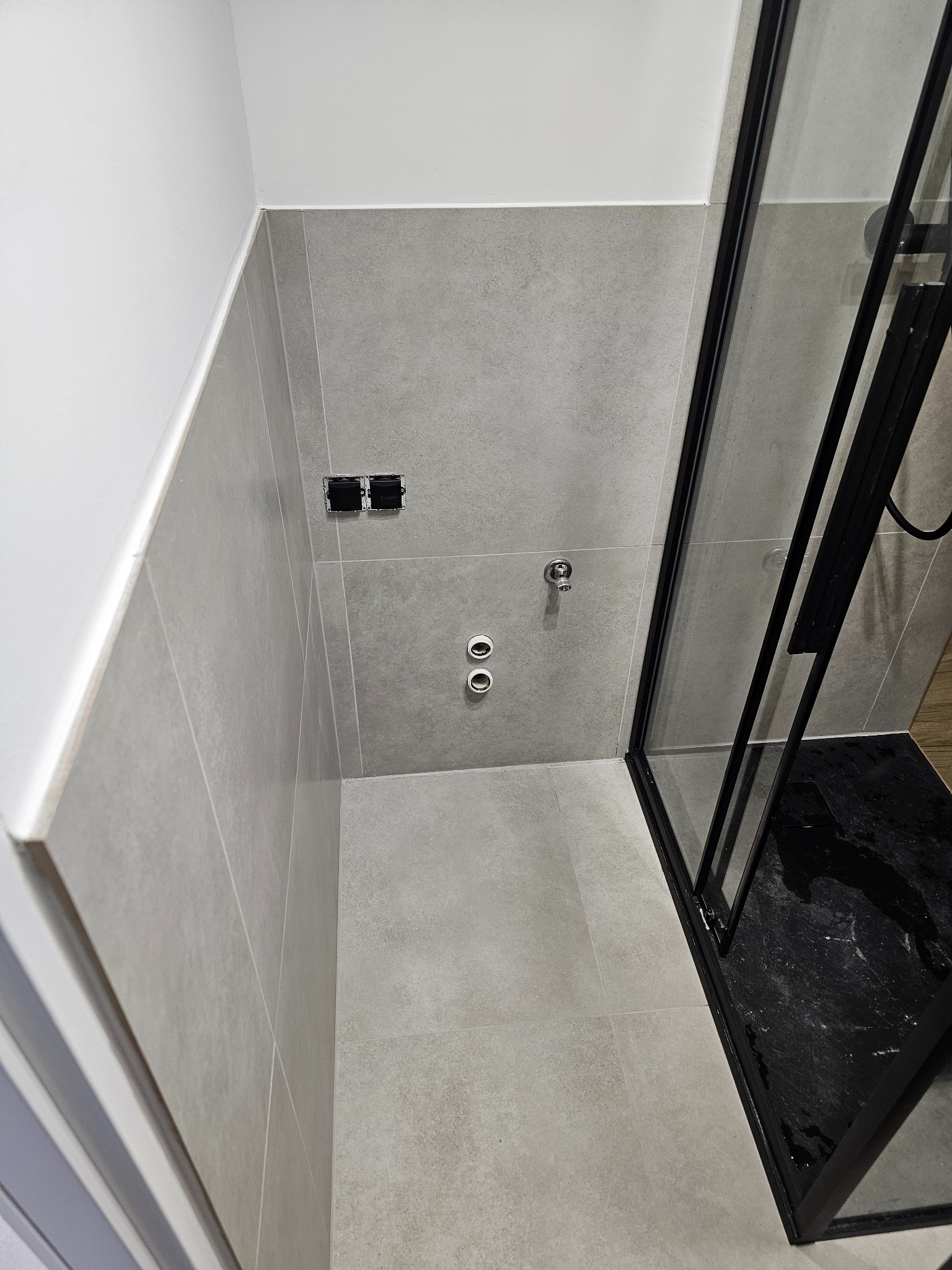 Wykończona łazienka z szarymi płytkami, czarną kabiną prysznicową i przygotowanymi przyłączami wodno-kanalizacyjnymi oraz elektrycznymi.